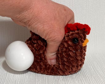 May include: A brown crocheted chicken with a red and orange comb and beak, holding a white ceramic egg.