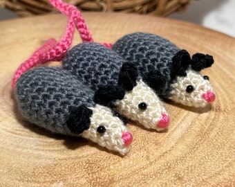 May include: Three crocheted gray and white opossums with pink noses. The opossums are connected by a pink yarn tail.