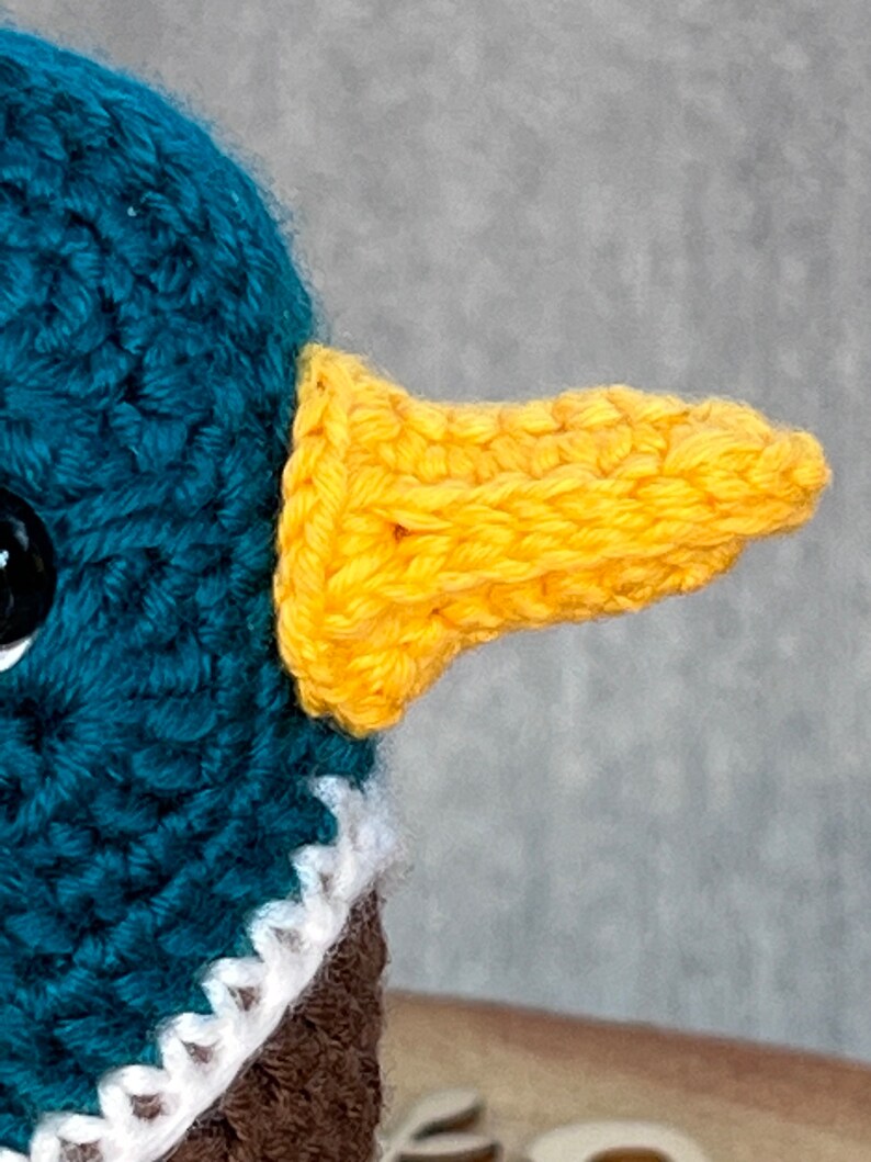 May include: Close-up of a crocheted duck toy with a teal head and a yellow beak. The duck is wearing a white crocheted collar.