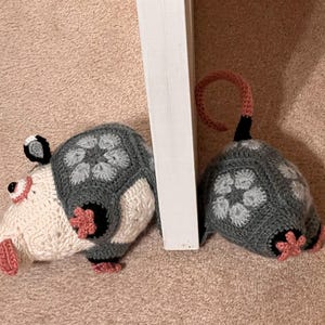 Könnte beinhalten: Zwei gehäkelte Stofftiere, eines weiß mit rosa Akzenten und das andere grau mit weißen Akzenten, sind nebeneinander platziert. Das weiße Tier hat eine lange Schnauze und eine rosa Nase. Das graue Tier hat einen Schwanz, der um seinen Körper gewickelt ist.