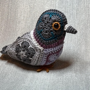 Op de afbeelding: Een handgehaakte duif in grijstinten, blauw, paars en wit. De vogel heeft een gedetailleerd ontwerp met een getextureerde uitstraling, een kleine snavel en oranje ogen. Een uniek decoratief item.