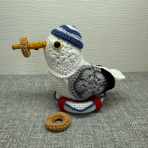 Könnte beinhalten: Handgefertigte Häkel-Möwenfigur mit blau-weiß gestreifter Mütze und Rettungsring. Die Möwe hält einen gelb-braunen Donut im Schnabel. Ein zweiter Donut liegt auf der Oberfläche.