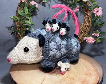 May include: A crocheted gray and white opossum with a pink tail, holding three smaller crocheted opossums on its back. The opossums are sitting on a wooden log with a floral wreath behind them.