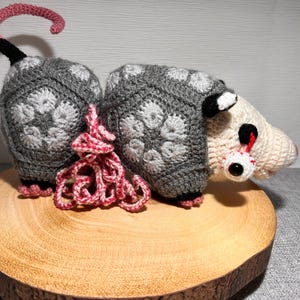 Puede incluir: Un juguete de peluche de opossum tejido a crochet con un cuerpo de patrón gris y blanco, una cola rosa y una cara blanca con un ojo grande de borde rojo. El opossum está sentado sobre una superficie de madera.