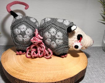May include: A crocheted opossum plush toy with a grey and white patterned body, pink tail, and white face with one large, red-rimmed eye. The opossum is sitting on a wooden surface.