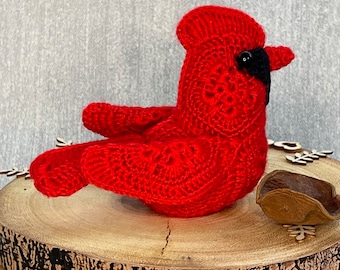 Puede incluir: Un pájaro cardenal rojo tejido a mano, con un ojo y un pico negros. El pájaro está posado sobre una rodaja de madera, mostrando su detallada artesanía. El color rojo vibrante y el intrincado patrón de ganchillo lo convierten en un objeto decorativo encantador.