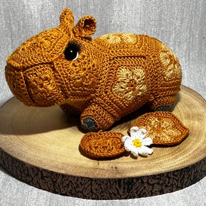Könnte beinhalten: Gehäkelte Capybara-Figur in Braun- und Beigetönen mit schwarzen Augen. Das handgefertigte Tier wird auf einer Holzscheibe präsentiert, mit zwei gehäkelten blattförmigen Teilen und einer kleinen weißen Blume.