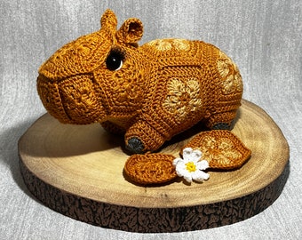 May include: A crocheted capybara figurine in shades of brown and tan, with black eyes. The handmade animal is displayed on a wooden slice, with two crocheted leaf-shaped pieces and a small white flower.
