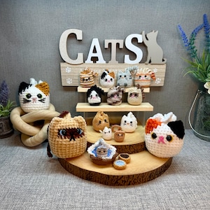 May include: A collection of crocheted cat figurines in various colors and poses, including a calico cat, a tabby cat, and a Siamese cat. The figurines are arranged on a wooden shelf with the word "CATS" displayed above.