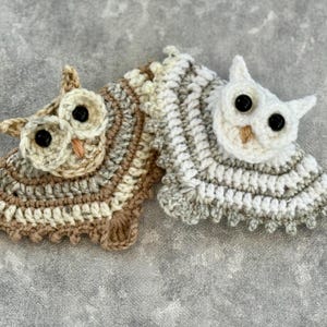 May include: Two crocheted owl ornaments. One owl is beige and brown, the other is white and gray. Both have black button eyes and small orange beaks. The owls are lying on a gray surface.