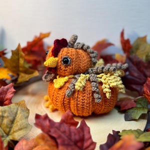 May include: A crocheted pumpkin with a rooster head and a brown, yellow, and orange color scheme. The pumpkin is surrounded by fall leaves.