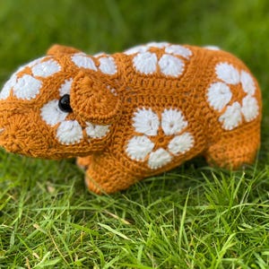 May include: A crocheted stuffed animal shaped like a guinea pig. The guinea pig is brown with white flower-shaped accents. It is sitting on a bed of green grass.