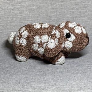 May include: A crocheted bunny toy in brown and white. The bunny features large black eyes and a small white tail. The body is brown with white floral patterns. The ears and feet are white. This soft toy is a charming, handmade item.