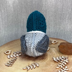 May include: A teal blue and grey knitted bird toy. The bird is sitting on a wooden surface with wooden leaves and a heart.