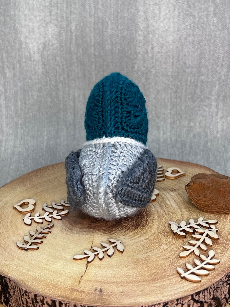 May include: A teal blue and grey knitted bird toy. The bird is sitting on a wooden surface with wooden leaves and a heart.