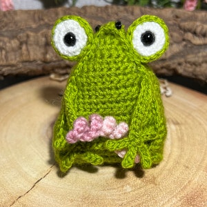 May include: A green crocheted frog with white eyes and black pupils. The frog is sitting on a wooden surface and has pink crocheted details on its feet.