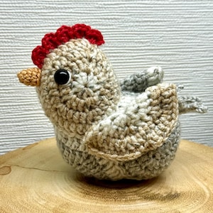 Pode incluir: Um galo de crochê feito à mão com crista vermelha, olhos pretos e bico castanho. O corpo é feito de uma mistura de fios bege, cinza e branco. As asas são de um tom mais claro das mesmas cores. O galo está sobre uma superfície de madeira.