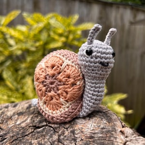 Może przedstawiać: Ręcznie robiony ślimak z włóczki, z szarym ciałem i różowo-beżową muszlą. Ślimak siedzi na pniu drzewa.