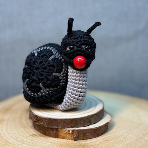 May include: A black and gray crocheted snail with a red nose. The snail is sitting on a wooden platform.