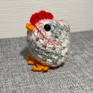 May include: A handmade crochet chicken figurine. The chicken is primarily gray, pink, and white, with a red comb, orange beak, and orange feet. The eyes are black. The figurine is sitting on a gray textured surface.