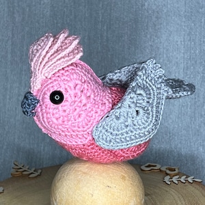 Puede incluir: Un pájaro de crochet rosa y gris con ojos negros, posado sobre una bola de madera. El pájaro tiene una cresta rosa y alas grises.