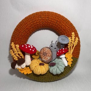Peut inclure: Une couronne d'automne en crochet avec une base marron et des accents verts. La couronne est décorée de champignons, de citrouilles et d'un escargot en crochet.