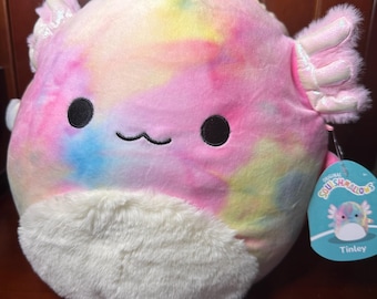 Puede incluir: Un peluche de ajolote con un patrón de teñido anudado en rosa, amarillo y azul. Tiene ojos negros, una boca pequeña y branquias esponjosas e iridiscentes. El juguete tiene una barriga blanca y esponjosa y una etiqueta que dice "Tinley".