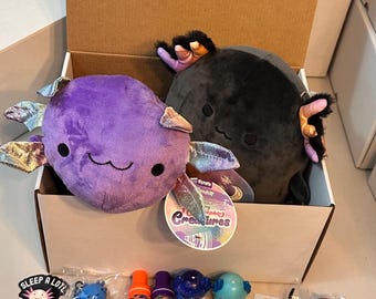 Puede incluir: Una caja de cartón llena de peluches de ajolotes y varios productos con temática de ajolotes. Los peluches son morados y negros. La mercancía incluye llaveros, sellos, pegatinas y una tarjeta navideña. La etiqueta dice "Squishmallows Creatures".