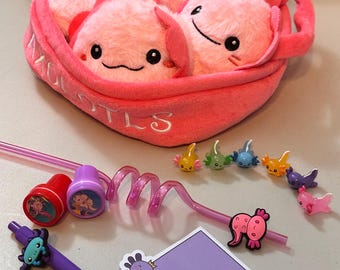 Puede incluir: Una cesta de peluche rosa con la palabra "AXOLOTLS" bordada en ella, llena de cinco peluches axolotl rosas. La cesta está rodeada de varios accesorios con temática de axolotl, incluyendo una pluma morada con un dije de axolotl, un bloc de notas morado con una calcomanía de axolotl, dos sellos de axolotl rojo y morado, una pajita rosa y ocho pequeñas figuras de axolotl.
