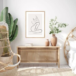 May include: A framed minimalist art print featuring a continuous line drawing of a seated figure. The artwork is in a natural wood frame and hangs above a wooden cabinet. The room is decorated in neutral tones with plants and a hanging chair.