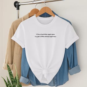 Puede incluir: Camiseta blanca con el texto "I'll be a bad bitch again soon. I'm just a little anxious right now." La camiseta est&aacute; anudada a la cintura y colgada de una percha de madera. Una chaqueta de pana color beige y una camisa vaquera est&aacute;n detr&aacute;s.