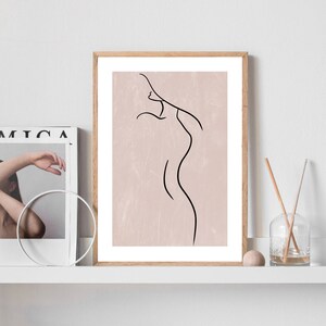 May include: A framed print with a minimalist line drawing of a person's body on a light pink background.