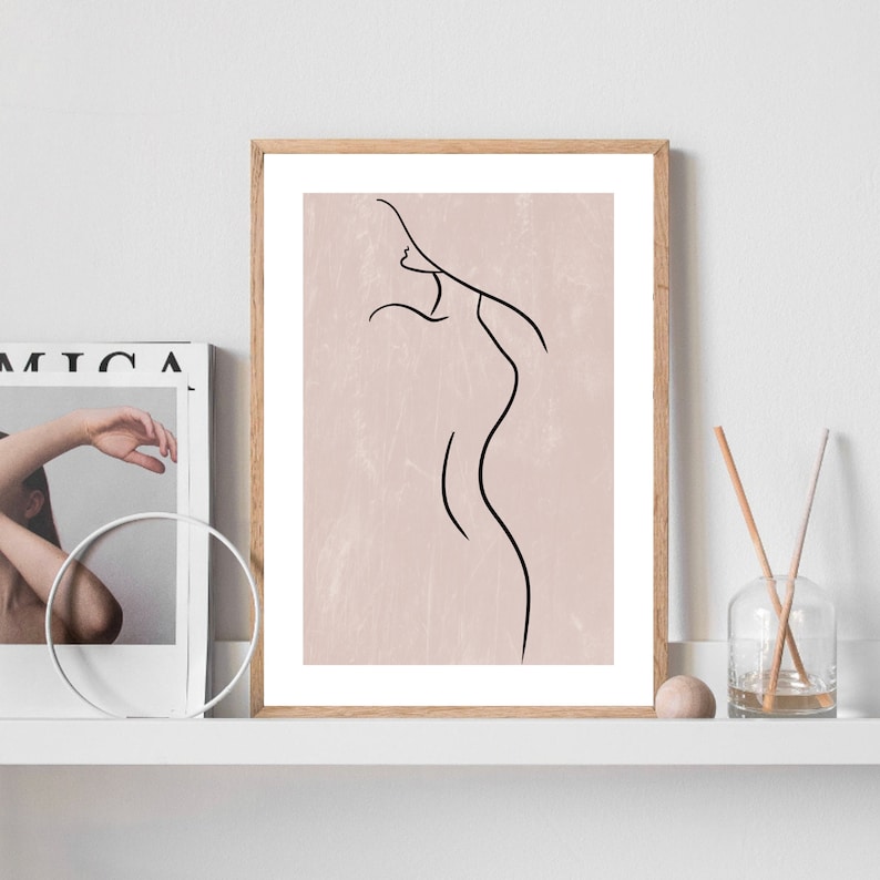 May include: A framed print with a minimalist line drawing of a person's body on a light pink background.