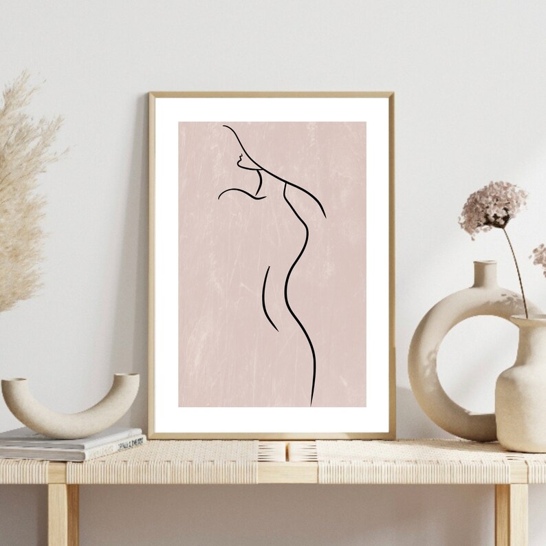 May include: A black line drawing of a person's body on a light pink background. The drawing is framed in a gold frame and is displayed on a shelf with other decor.