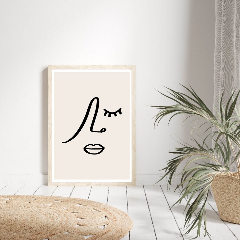 K&ouml;nnte beinhalten: Ein minimalistischer Line-Art-Druck eines Gesichts mit geschlossenem Auge, Nase und Mund. Der Druck ist gerahmt und hat einen beigefarbenen Hintergrund.