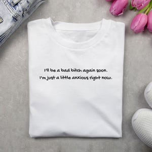 Puede incluir: Camiseta blanca con el texto "I'll be a bad bitch again soon. I'm just a little anxious right now." en letras negras. La camiseta est&aacute; doblada sobre una superficie gris claro, con unos vaqueros y zapatillas blancas.