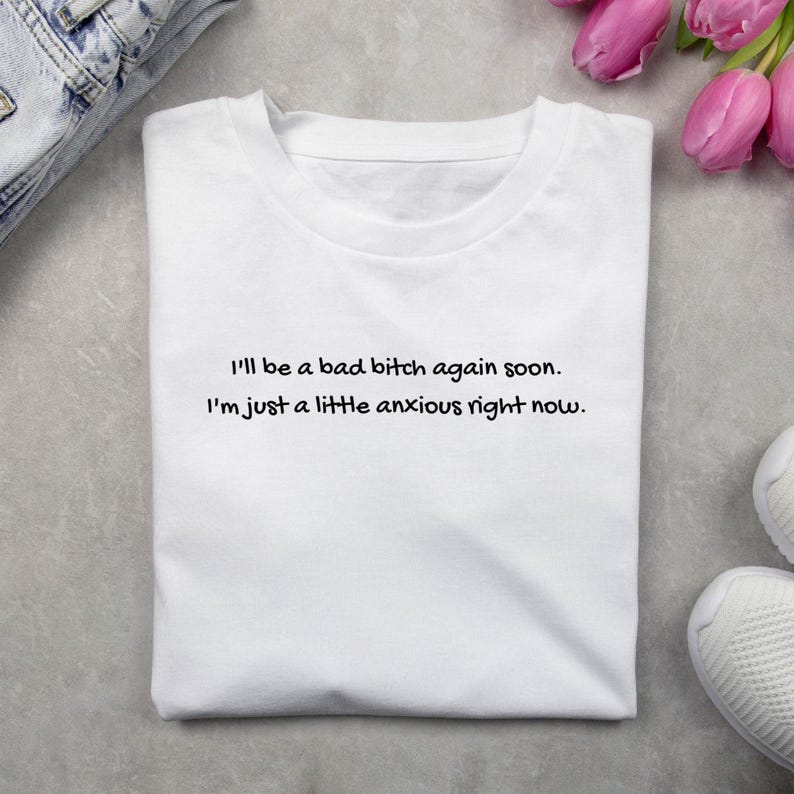 Puede incluir: Camiseta blanca con el texto "I'll be a bad bitch again soon. I'm just a little anxious right now." en letras negras. La camiseta est&aacute; doblada sobre una superficie gris claro, con unos vaqueros y zapatillas blancas.