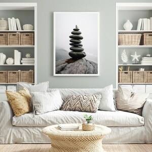 May include: A framed print of a stack of gray stones balanced on a piece of driftwood. The stones are stacked in a pyramid shape, with the top stone being the smallest. The background is a blurry image of a beach with a foggy sky.