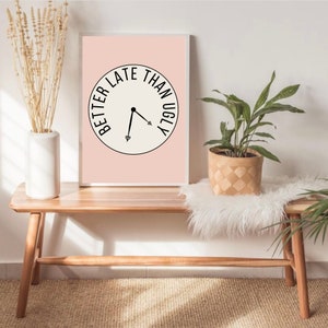 May include: A pink framed print with a white background and black text that reads "Better Late Than Ugly" in a circle with a clock hand pointing to the text. The print is sitting on a wooden bench with a white faux fur throw and a potted plant.