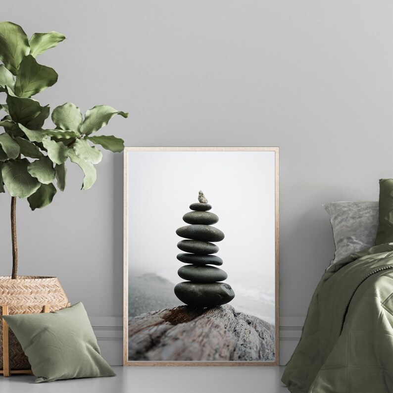May include: A stack of gray stones balanced on a weathered piece of driftwood, with a small white rock on top. The stones are arranged in a pyramid shape, and the driftwood is lying on a beach with a foggy background.