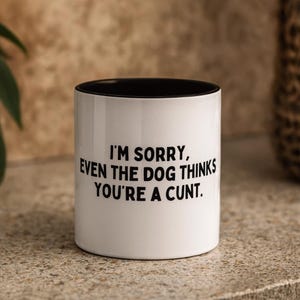 May include: White ceramic mug with a black interior. The mug features the text "I'M SORRY, EVEN THE DOG THINKS YOU'RE A CUNT" in black, centered on the front. The mug is set on a neutral-toned surface.