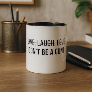 Puede incluir: Taza de cerámica blanca con interior negro, con el texto negro "LIVE, LAUGH, LOVE DON'T BE A CUNT". La taza está sobre una superficie de madera. Un bolígrafo y un cuaderno son visibles en el fondo, así como un portalápices.