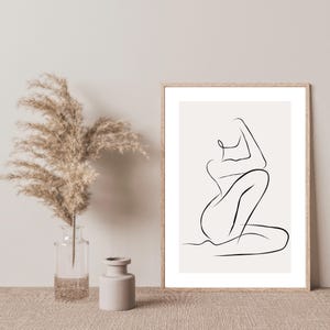 May include: A framed minimalist line art print of a seated figure in black on a cream background. The artwork is displayed next to a clear glass vase with dried pampas grass and a small beige ceramic vase.