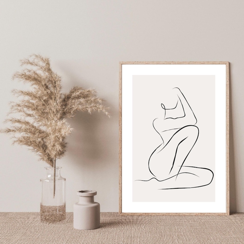 May include: A framed minimalist line art print of a seated figure in black on a cream background. The artwork is displayed next to a clear glass vase with dried pampas grass and a small beige ceramic vase.