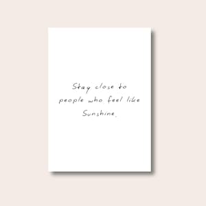 Puede incluir: Una tarjeta rectangular blanca con el texto manuscrito: "Stay close to people who feel like Sunshine." El texto está en cursiva negra. El fondo es de color beige claro.