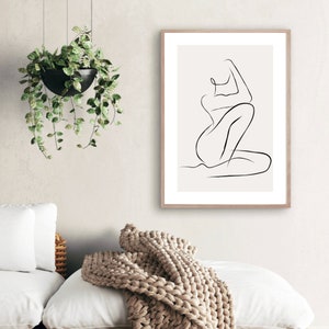 May include: A framed minimalist art print featuring a black line drawing of a seated figure. The artwork is set against a cream background and framed in light wood. The image is displayed in a well-lit room with a hanging plant and a cozy blanket.