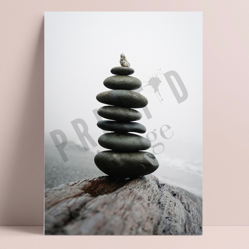 May include: A stack of gray stones balanced on top of a weathered wooden log. The stones are arranged in a pyramid shape, with a small white rock on top. The background is a blurry image of a foggy beach.