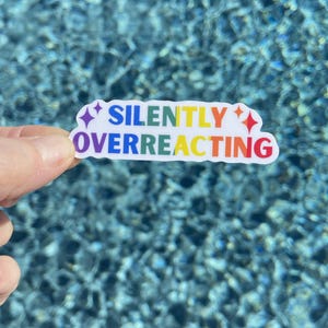 May include: A white sticker with the text "SILENTLY OVERREACTING" in rainbow colors. The sticker has a star on each end and is held against a background of rippling blue water.