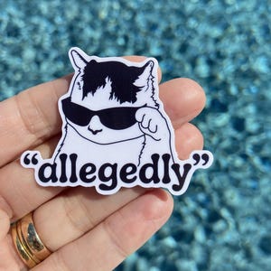 May include: A white sticker featuring a cartoon cat with black and white markings wearing sunglasses. The word "allegedly" is written in black font below the cat. The sticker is held in a hand.