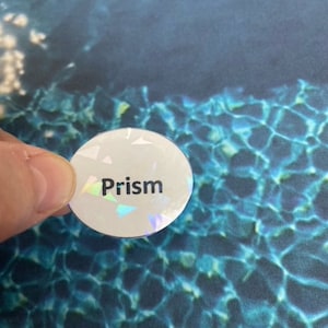 May include: A round, white sticker with the word "Prism" printed in black. The sticker has a holographic, iridescent finish. The background is a blue and teal water pattern.